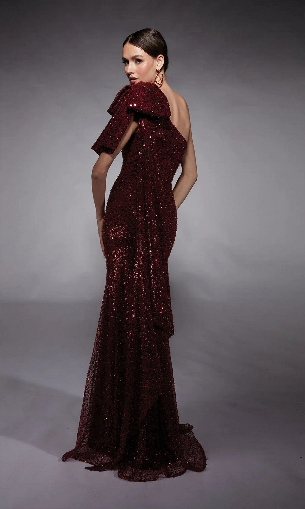 A woman poses in the Alyce 27771, a floor-length burgundy formal dress with a ruffled off-the-shoulder sleeve, against a plain gray background.